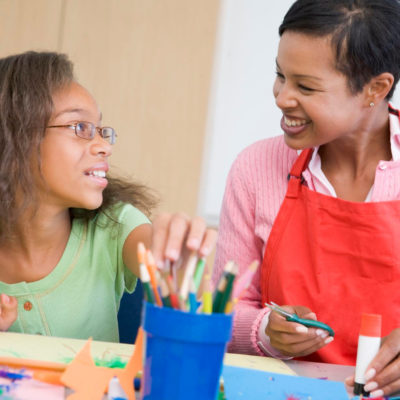 Teacher working with student on an art project at a table with markers and crayons