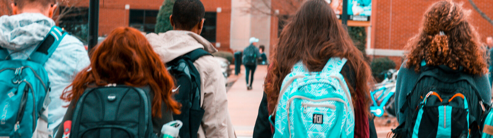 Students walking into school, image from behind so you can only see back of their heads and backpacks